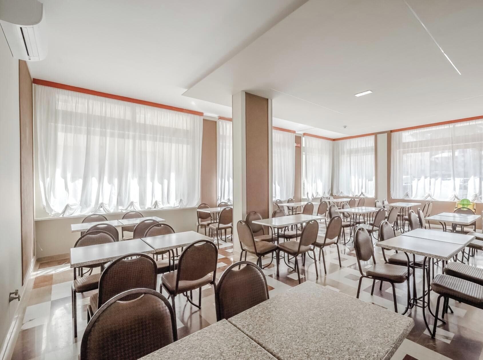 Un comedor de hotel con varias mesas y sillas, ventanas amplias con cortinas y piso de baldosas.