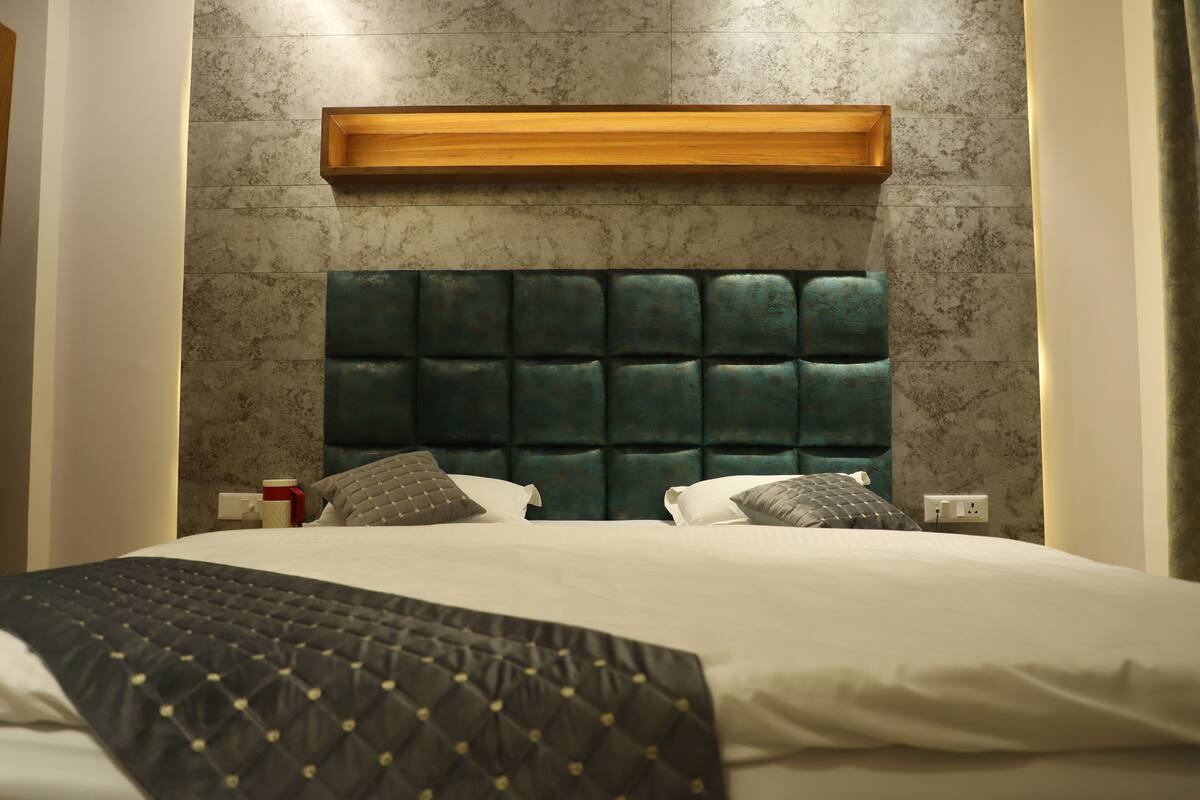A neatly made bed with a quilted headboard, a bedside table with a red cup, and a power outlet on the wall.