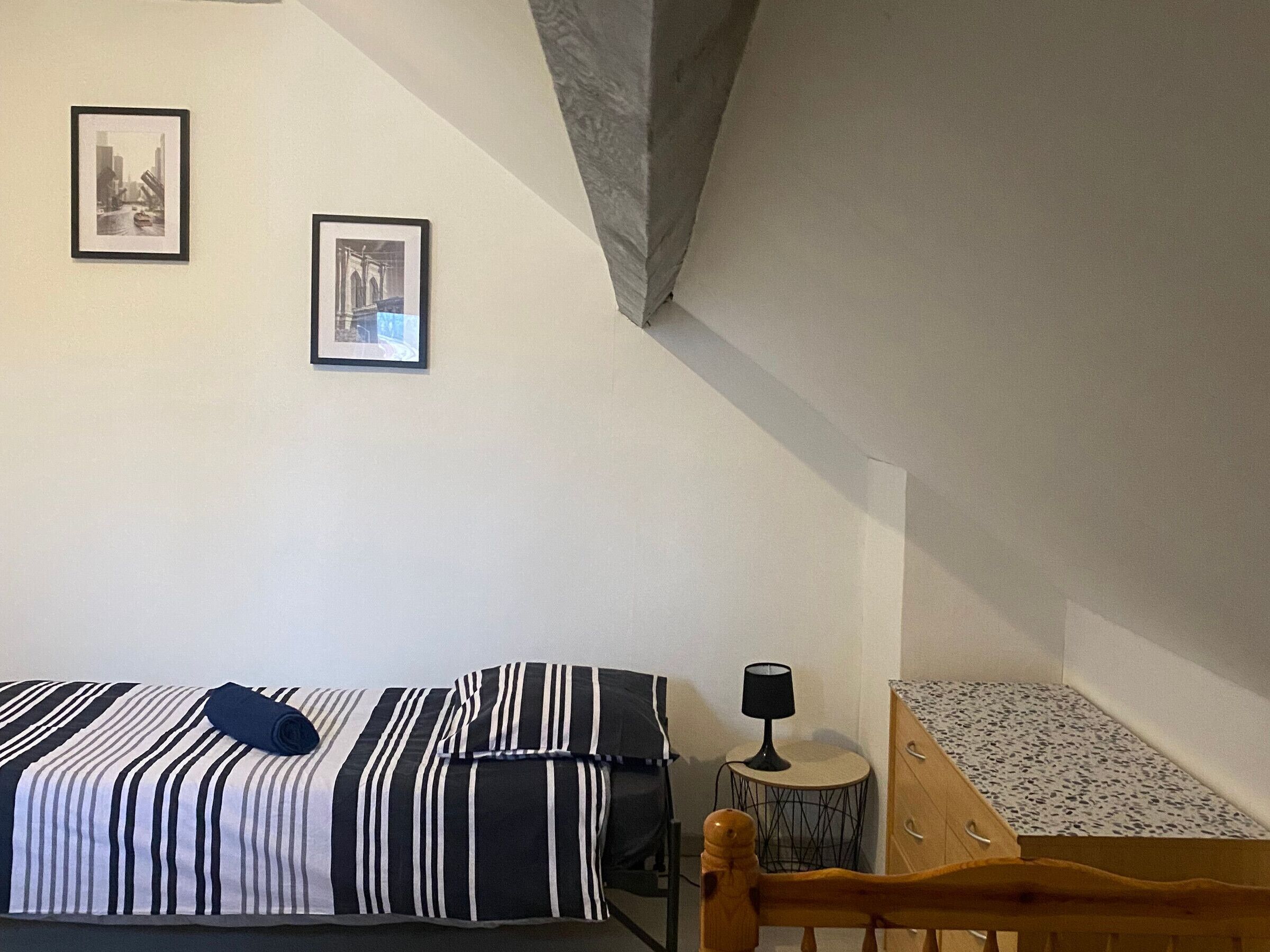 A bedroom with a bed, a small round table with a lamp, and two framed pictures on the wall.