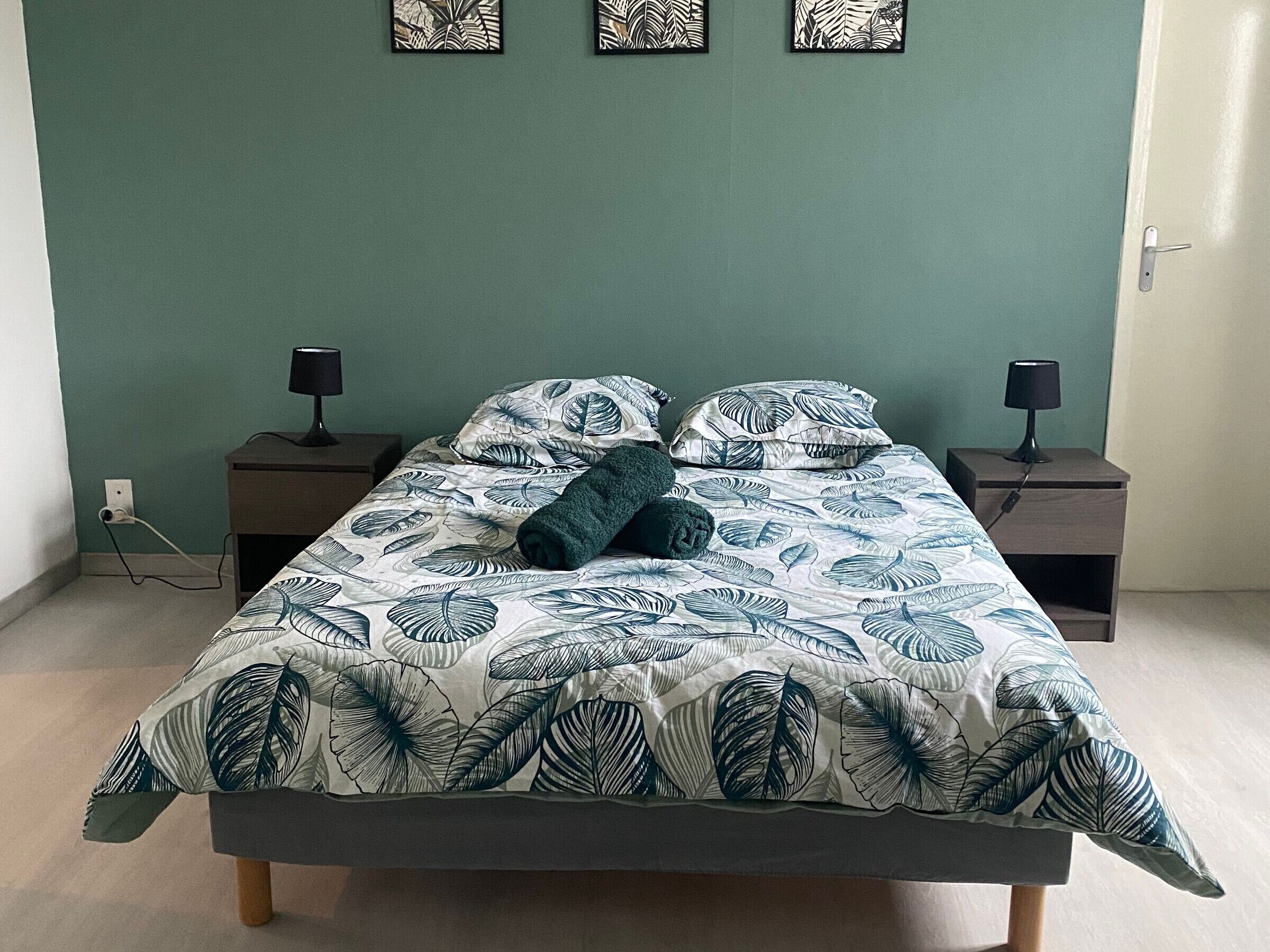 Un lit recouvert d’une couvre-lit à motifs de feuilles, deux tables de chevet avec des lampes et un mur orné de trois œuvres d’art encadrées.