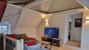 A modern loft-style living room with a sloped ceiling, a grey sofa with red and brown cushions, a flat-screen TV, and a wooden coffee table. - Renovated house in the heart of Groix, 9 sleeps (Groix)