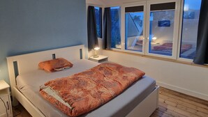 A bedroom with a bed, a nightstand, a lamp, and a window with curtains. - Renovated house in the heart of Groix, 9 sleeps (Groix)