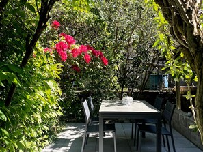 Uma área de refeições ao ar livre com mesa e cadeiras, cercada por vegetação exuberante e uma árvore com flores cor-de-rosa.