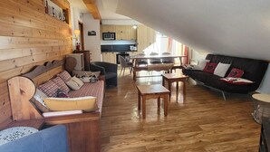 A cozy living room with a wooden sofa, a blue armchair, and a black sofa with heart-patterned cushions. A wooden dining table with chairs is in the background. - Spacious apt for 6, pool & sauna, near slopes & shops (Les Orres)