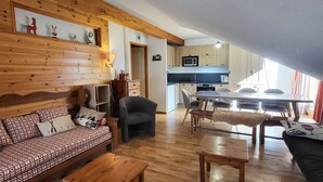 A cozy living area with a wooden sofa, a coffee table, and a dining table with chairs. A kitchen with a microwave and cabinets is visible in the background. - Spacious apt for 6, pool & sauna, near slopes & shops (Les Orres)