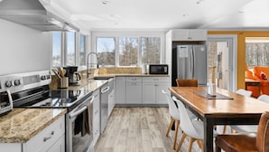 Une cuisine moderne équipée d’un four, d’un micro-ondes et d’un réfrigérateur intégrés. Elle comprend une table et des chaises de salle à manger en bois, une grande fenêtre donnant sur les arbres et un intérieur lumineux et bien éclairé.