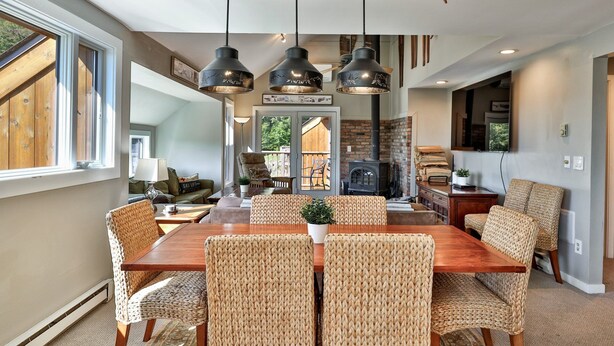 Un coin repas avec une table en bois et des chaises en osier, des poutres en bois apparentes au plafond et une cheminée visible au fond.