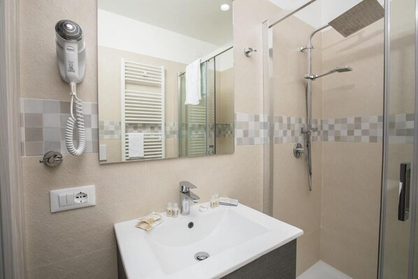 A modern bathroom with a glass shower enclosure, a hairdryer mounted on the wall, and a sink with toiletries. - Hotiday Vieste Lungomare  (Vieste)