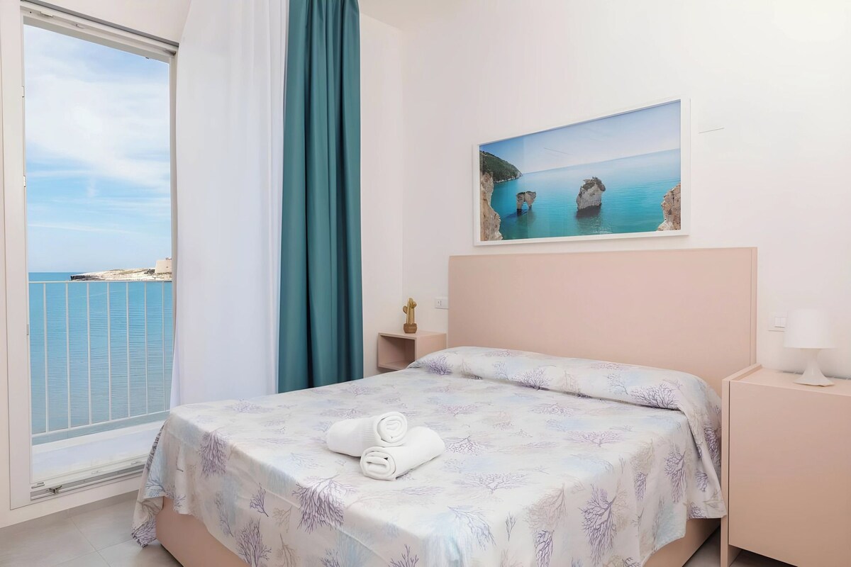 A bedroom with a bed, a nightstand, a painting of a coastal scene, and a view of the sea through a window.