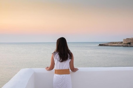 A person standing on a balcony overlooking a calm sea with a sunset in the background.