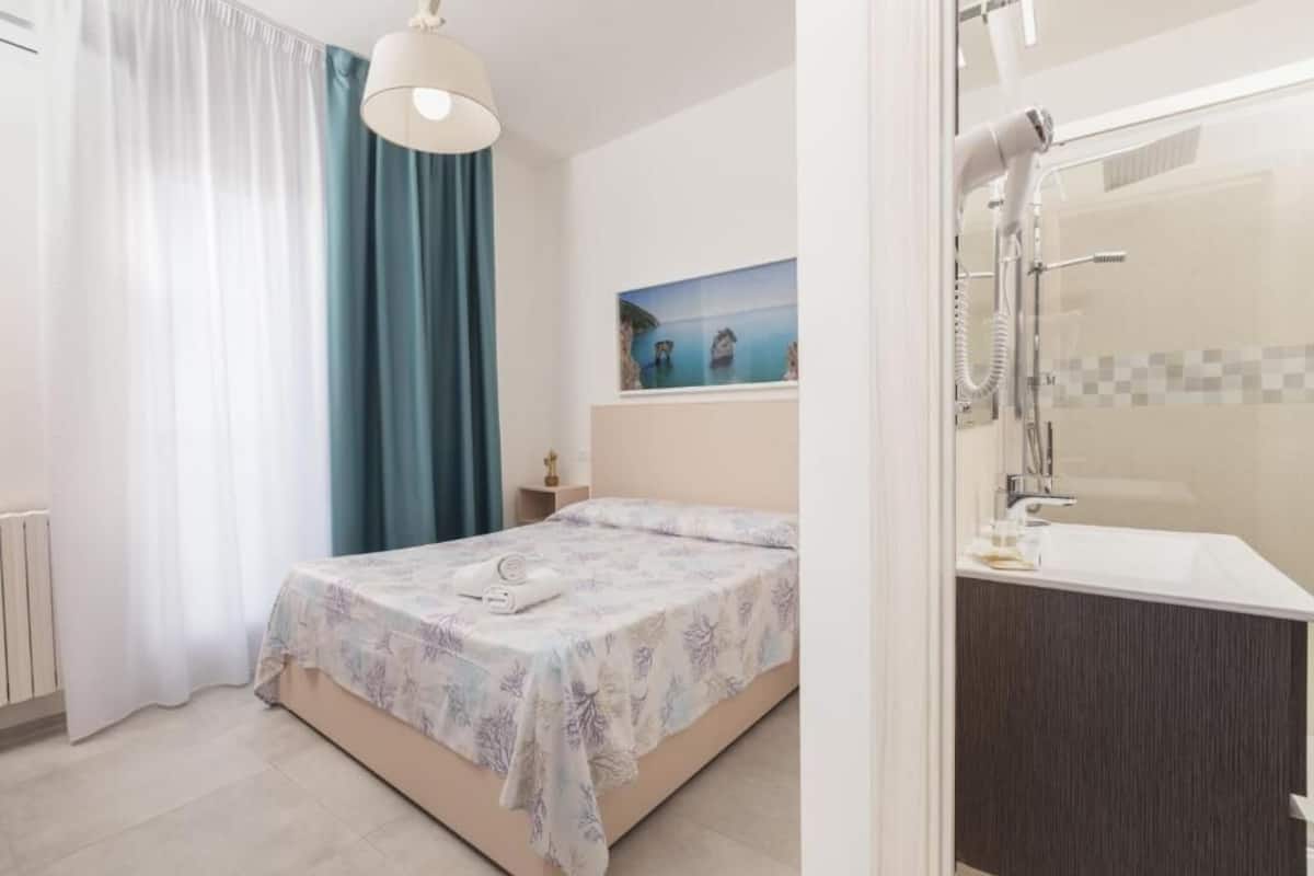 A bedroom with a bed, a nightstand, a painting, and a bathroom with a sink and mirror.