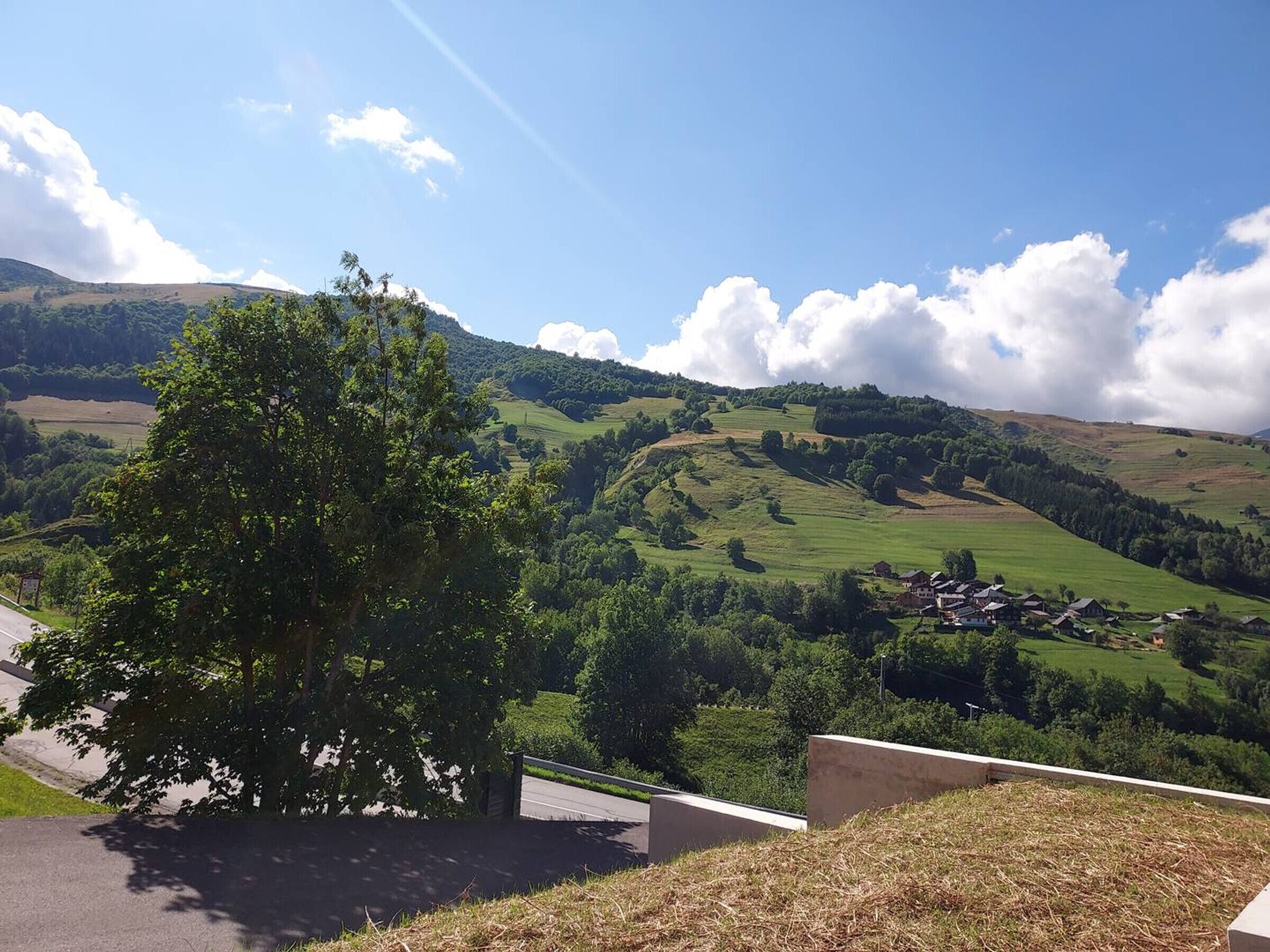 Ländliche Landschaft mit sanften Hügeln, einem Baum im Vordergrund und einem kleinen Dorf in der Ferne.