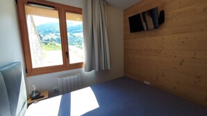 Una habitación con cama, ventana con cortinas, radiador y televisor montado en la pared.