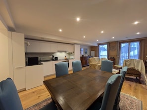 A modern kitchen with white cabinets, a dining area with a wooden table and blue chairs, and a view of the outdoors through large windows.