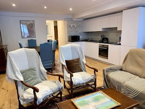 Una sala de estar moderna con dos sillones, un sofá, una mesa de centro y una zona de cocina con armarios y electrodomésticos blancos.