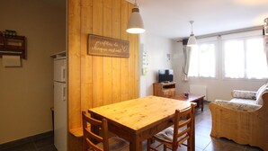 Une cuisine chaleureuse avec des murs en bois, une table à manger et des chaises. On peut voir un panneau sur le mur, un réfrigérateur et une télévision à l’arrière-plan.