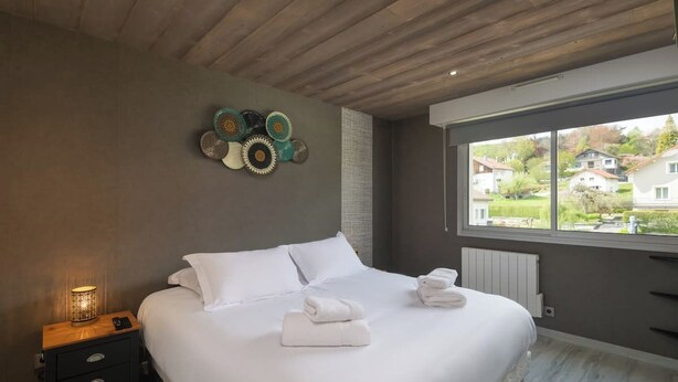 A bedroom with a large bed, a nightstand with a lamp, and a view of houses through a window. - Apartment "Maya" - 2-4 guests - Lake view, near the centre (Gérardmer)