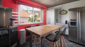 A modern kitchen with a wooden dining table, black chairs, a stainless steel refrigerator, and a window with a view of houses. - Apartment "Maya" - 2-4 guests - Lake view, near the centre (Gérardmer)