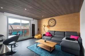 A modern living room with a grey sofa, a wooden coffee table, and a large window overlooking a balcony. - Apartment "Maya" - 2-4 guests - Lake view, near the centre (Gérardmer)