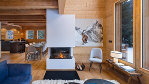 Interior acogedor de una cabaña con chimenea, paredes de madera, zona de comedor y un ventanal con vistas al exterior.