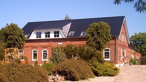 A large, red brick building with a dark tiled roof, surrounded by well-maintained hedges and a gravel driveway. - Ferienhof Eckhoff apartment no. 2 (Fehmarn)
