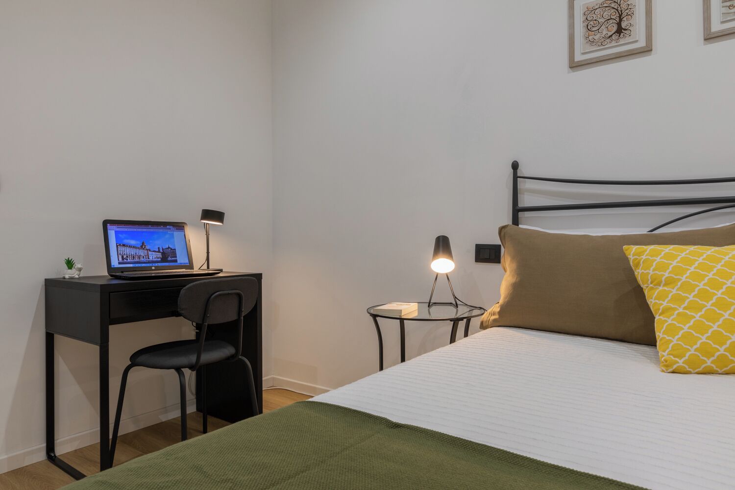 A neatly arranged bedroom with a bed, a desk with a laptop, a chair, a bedside table with a lamp, and a wall-mounted shelf.