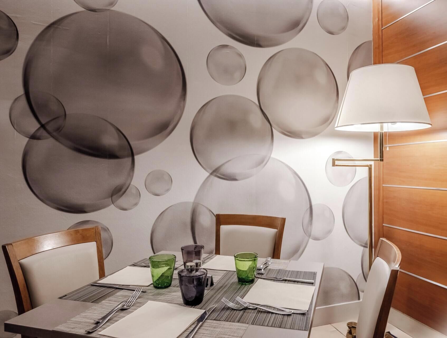 A dining area with a table set for two, a wall with a bubble pattern, and a lamp on the right.
