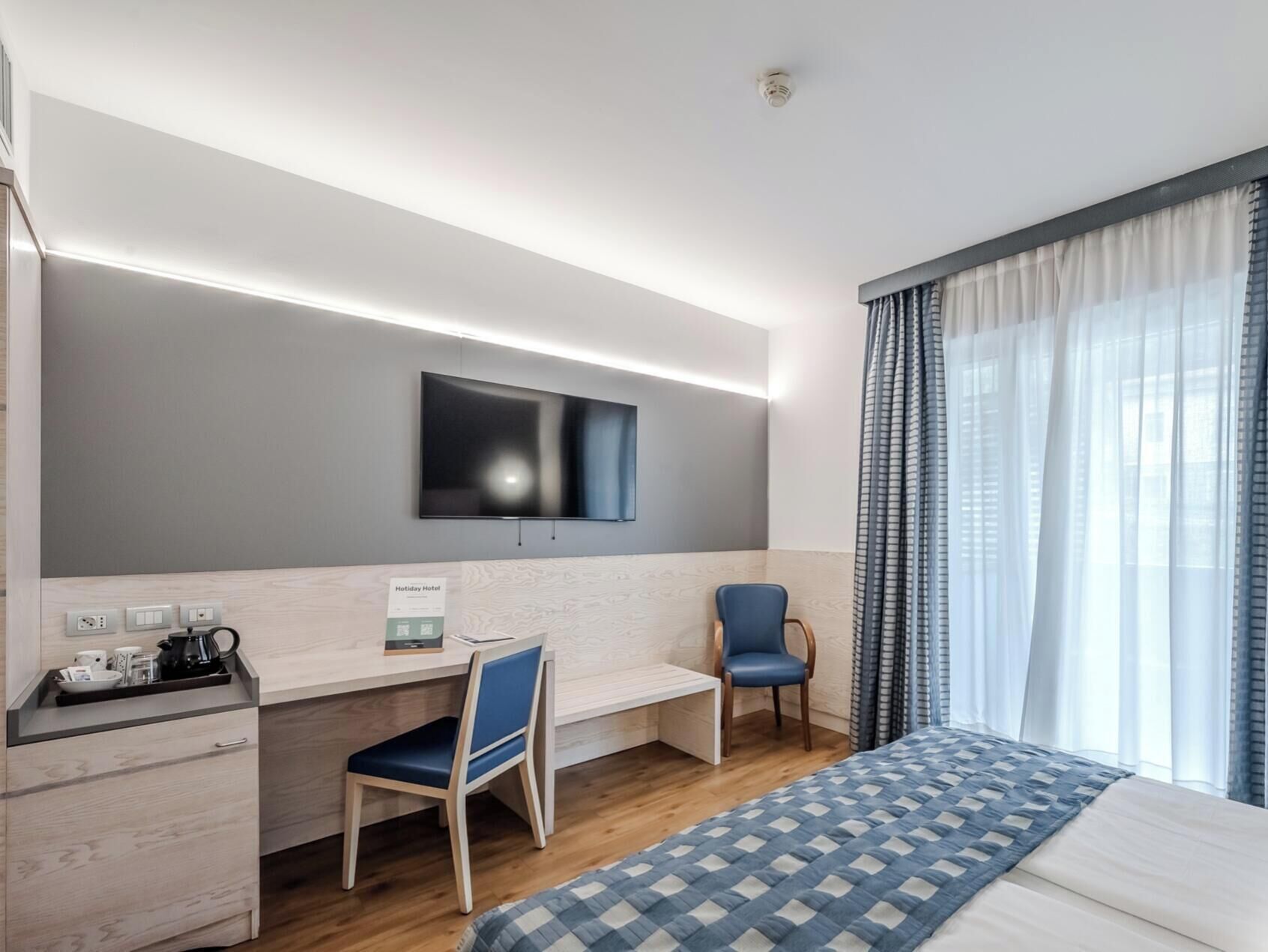 Une chambre d’hôtel équipée d’un lit, d’un bureau, d’une chaise, d’un téléviseur fixé au mur et d’une fenêtre avec des rideaux.