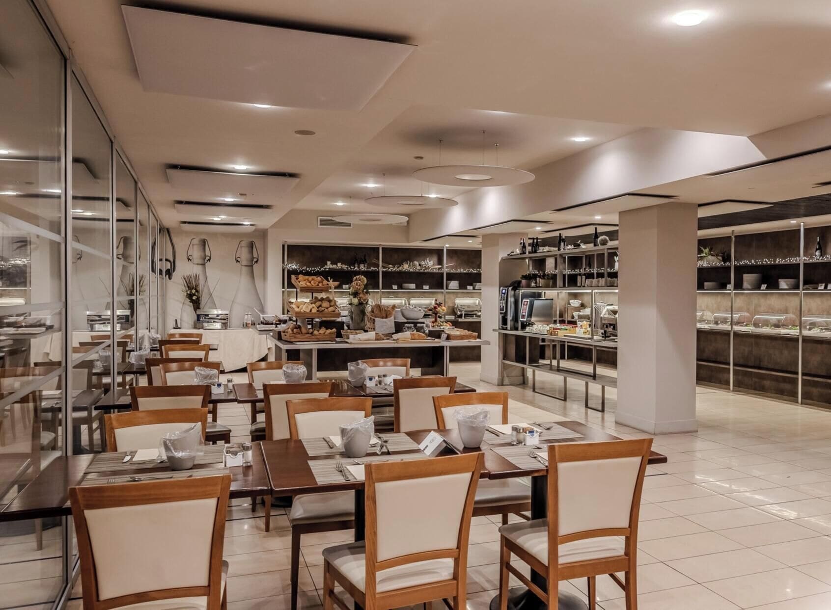 A modern dining area with tables and chairs, a buffet with various food items, and shelves with glassware.