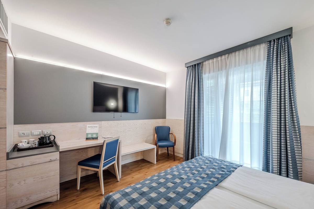 A hotel room with a bed, a desk, a chair, a TV mounted on the wall, and a window with curtains.