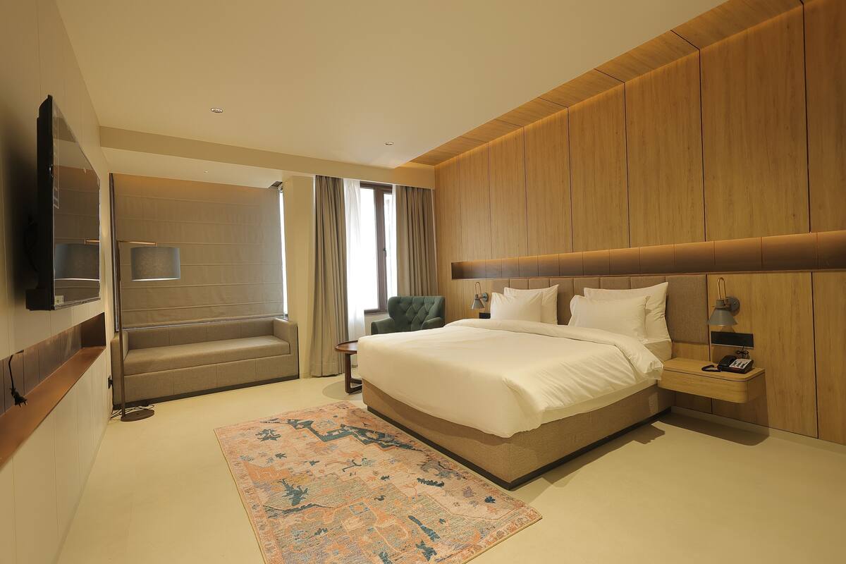 Una camera d'albergo moderna con un letto grande, un'area salotto con una poltrona, una TV a schermo piatto e un pannello parete in legno.