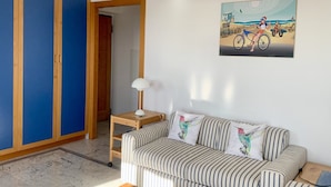 Un salon avec un canapé à rayures, un meuble en bois, une lampe et un tableau représentant une scène de plage.