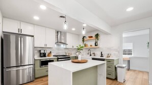 A modern kitchen with white cabinets, stainless steel appliances, a central island, and wooden flooring. - Ivy House / 4BR Boho Retreat Near UTD (Richardson)