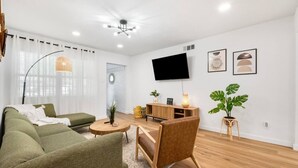 A modern living room with a sofa, a coffee table, a TV, and a plant. - Ivy House / 4BR Boho Retreat Near UTD (Richardson)