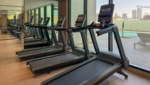 Een rij loopbanden in een sportschool met grote ramen en uitzicht op een stadsgezicht.
