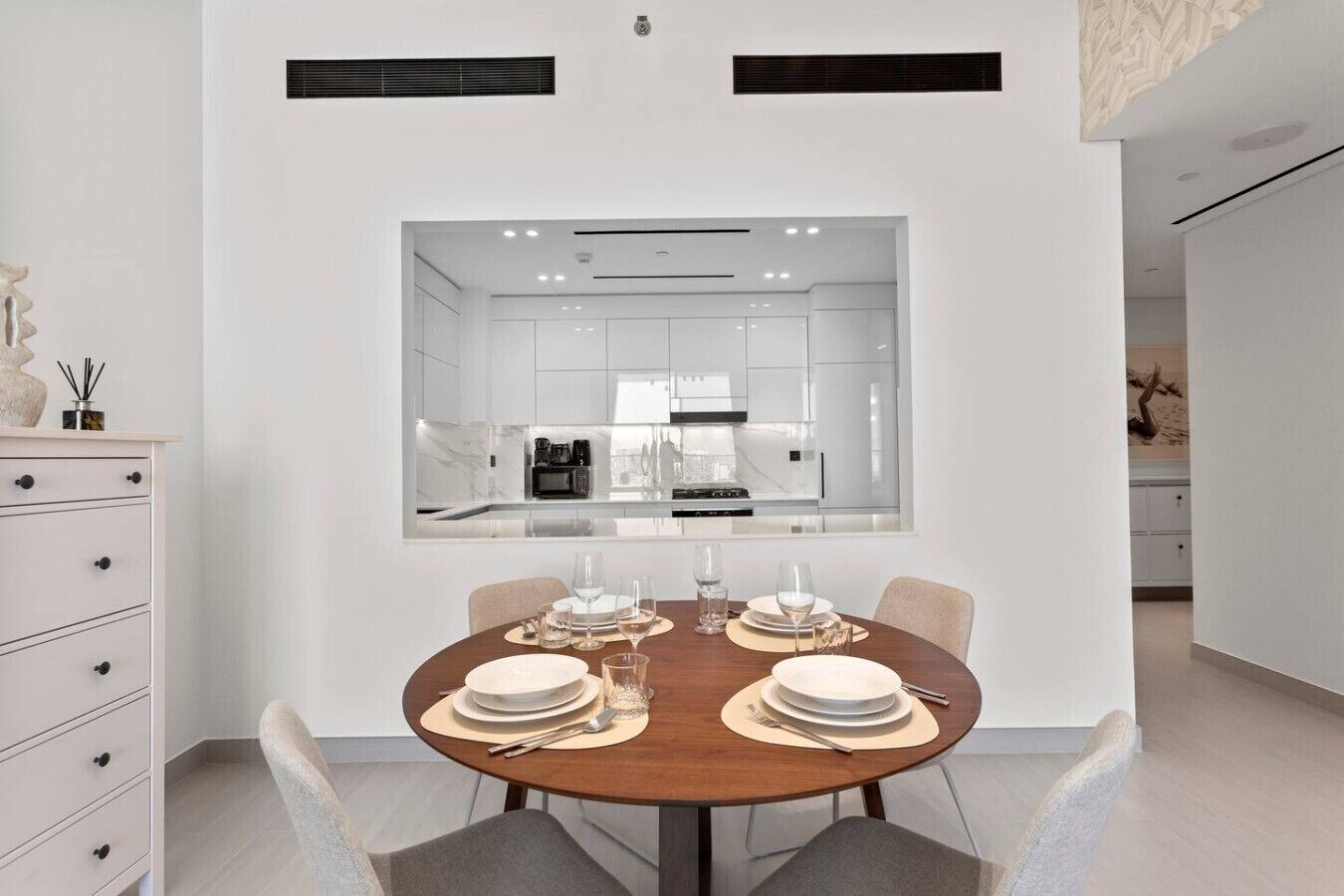 A modern dining area with a round table set for four, a kitchen visible through a glass partition, and a minimalist decor style.