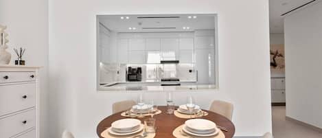 A modern dining area with a round table set for four, a kitchen visible through a glass partition, and a minimalist decor style.