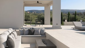A modern outdoor living area with a sofa and chairs, a ceiling fan, and a view of a hilly landscape.