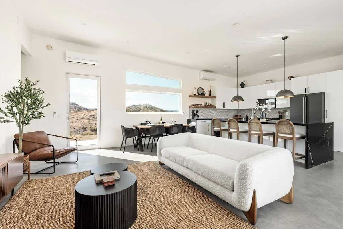 A modern living room with a white sofa, a dining area with a table and chairs, and a kitchen with a black refrigerator and pendant lights.