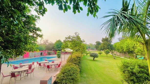 A swimming pool with red chairs and tables, surrounded by greenery and a well-maintained lawn. - Royal stay farm house (Gurugram)