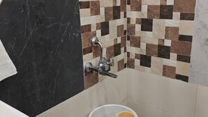 A bathroom with a black marble wall, a tiled shower area with a checkered pattern, a white sink, and a white toilet with a beige bucket. - Rawla Sarkar (Udaipur)