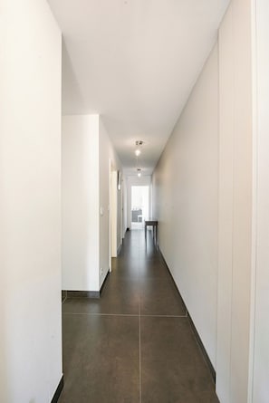 A long, narrow hallway with a dark floor and white walls, leading to a room with a table and chairs.