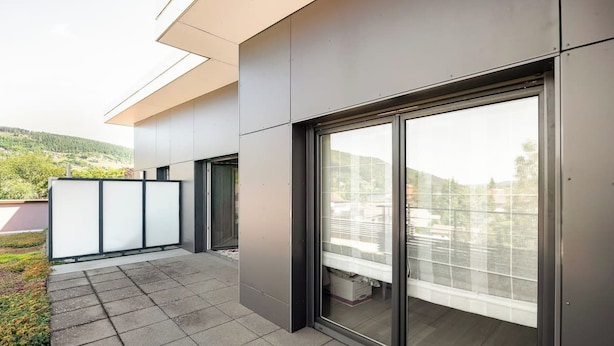 Een modern gebouw met een balkon, grote schuiframen en uitzicht op groen.