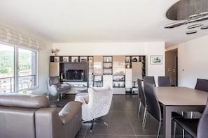 A modern living room with a sofa, armchair, and dining table. There is a television and a bookshelf with various items.