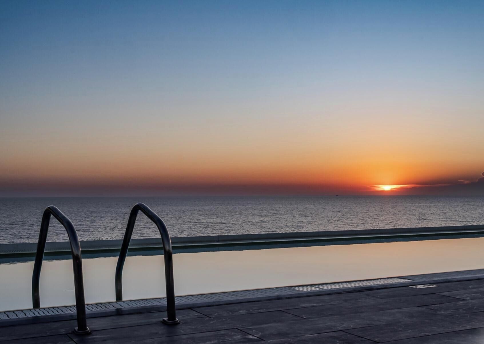 Blick auf ein Schwimmbecken mit Sonnenuntergang über dem Meer, Metallgeländern und einem wolkenlosen Himmel.