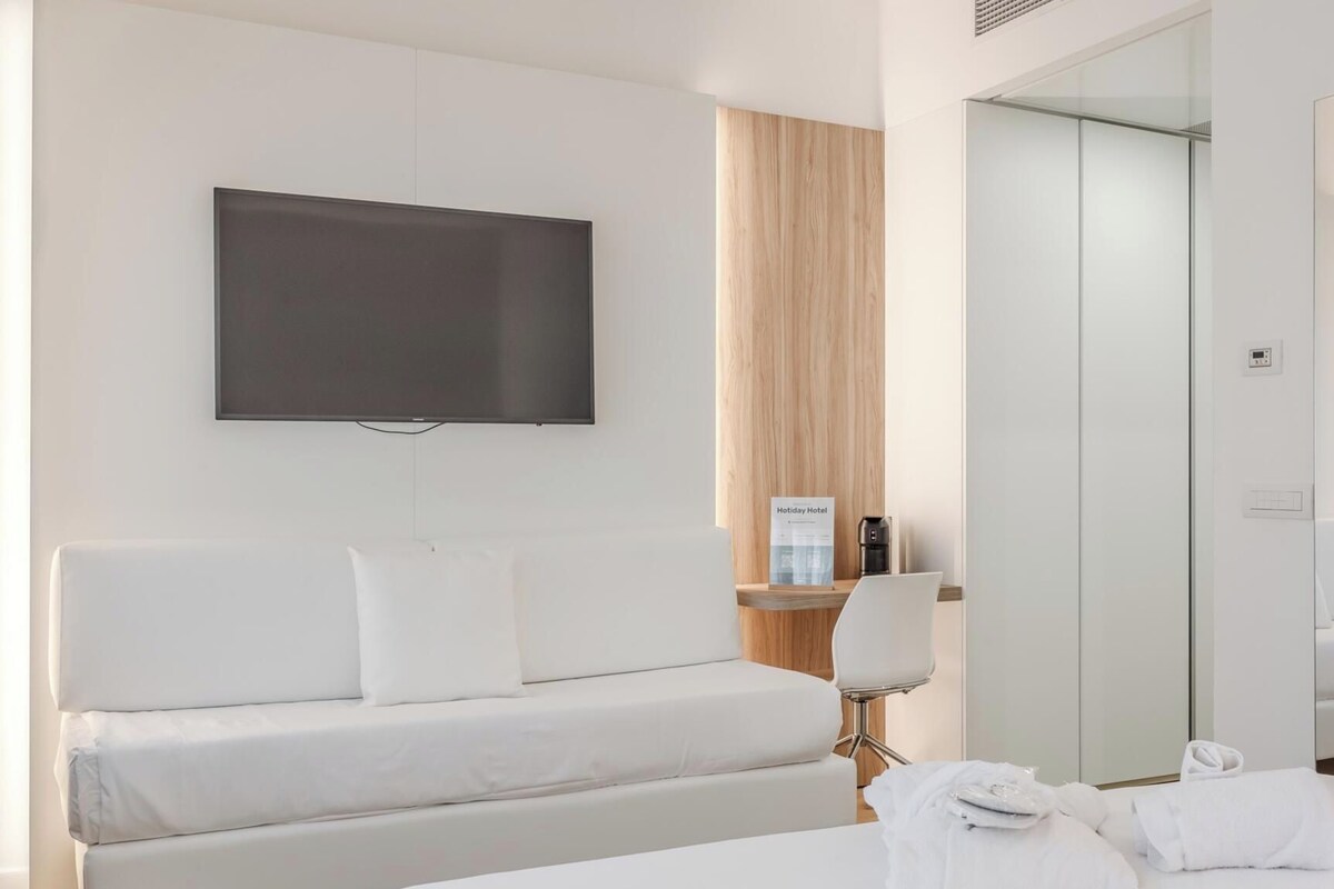 A modern hotel room with a large bed, a flat-screen TV mounted on the wall, a wooden accent wall, a small table with a coffee maker, and a mirror.