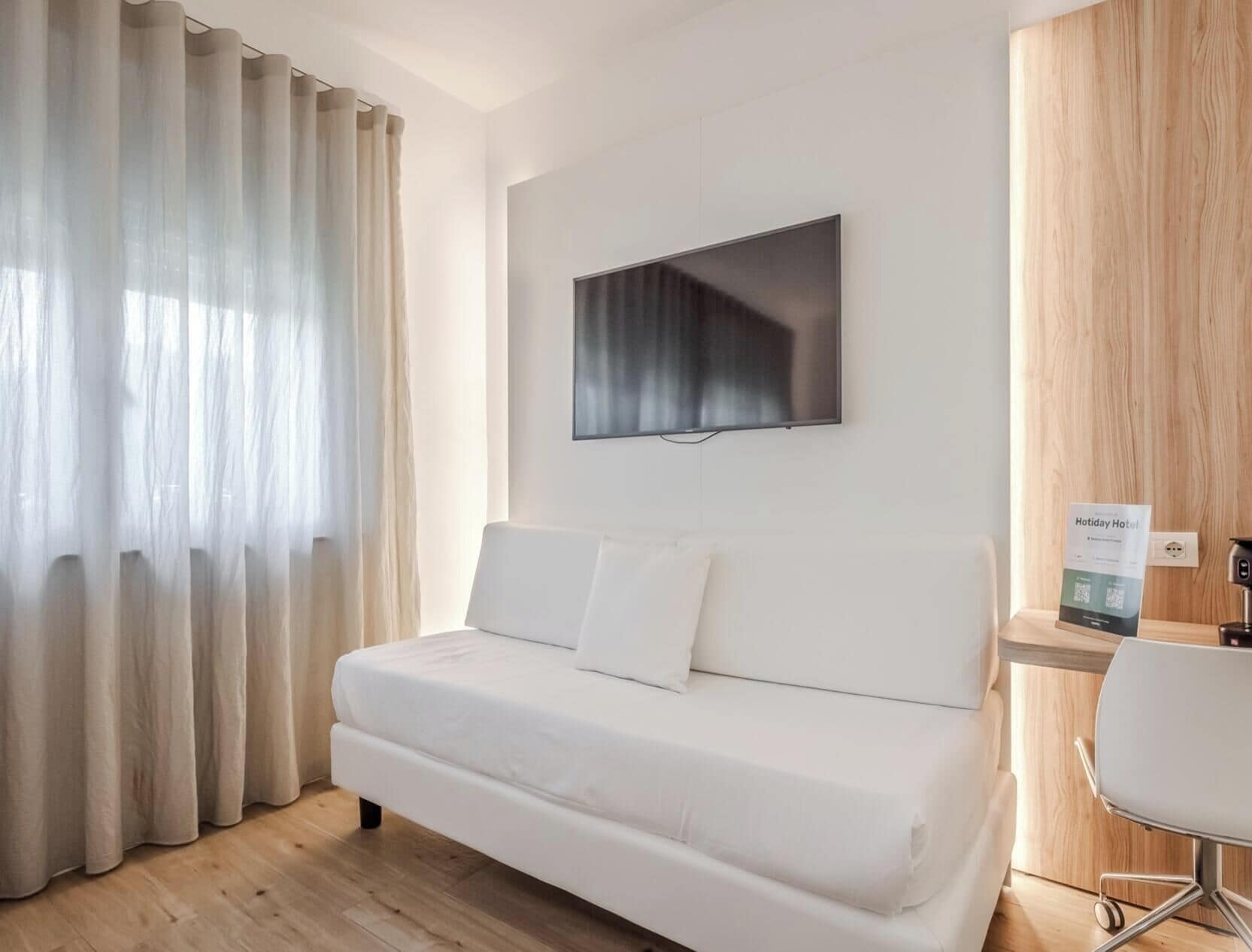 Una moderna camera d'albergo con un divano bianco, una TV a schermo piatto montata e una scrivania in legno con una sedia.