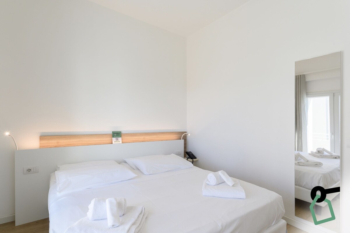 Ett hotellrum med en stor säng, ett sängbord, en liten avlastningsyta, en spegel och ett fönster med gardiner.