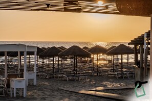 A beach with thatched umbrellas and sun loungers, a wooden deck, and a view of the ocean at sunset.
