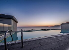 Zona de piscina con vista al océano al atardecer, terraza de madera y edificios modernos.